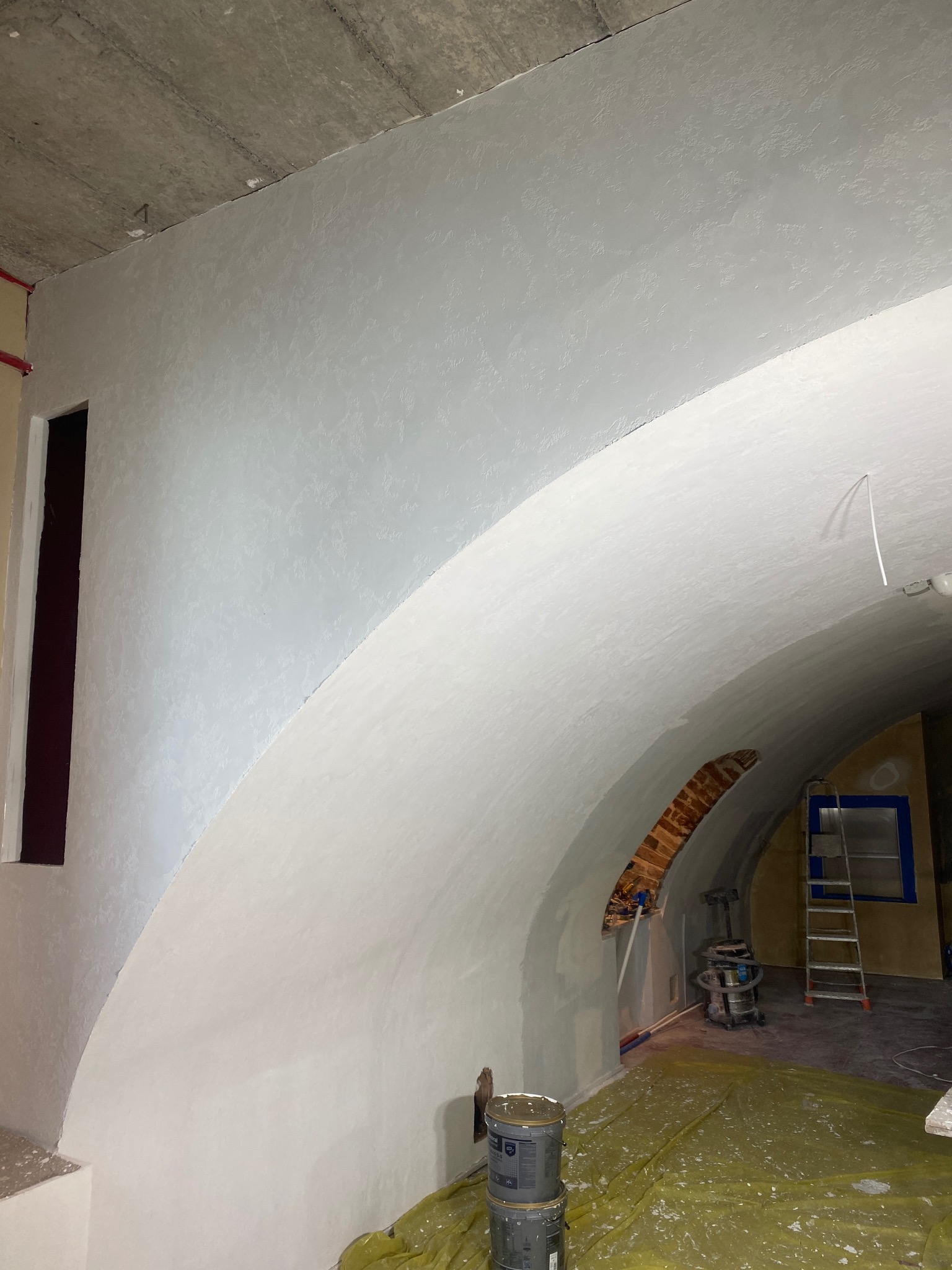 Wnętrze z łukowym sufitem po nałożeniu gładzi, widoczne wiaderka z farbą, drabina i zakryta folią podłoga.