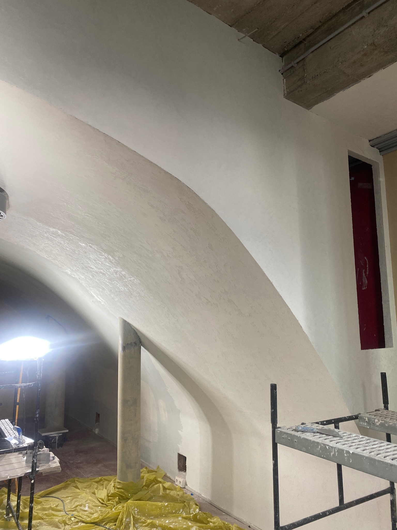 Wnętrze w trakcie prac wykończeniowych, widoczna gładź na łukowym suficie, rusztowanie i lampa budowlana, podłoga zabezpieczona folią.