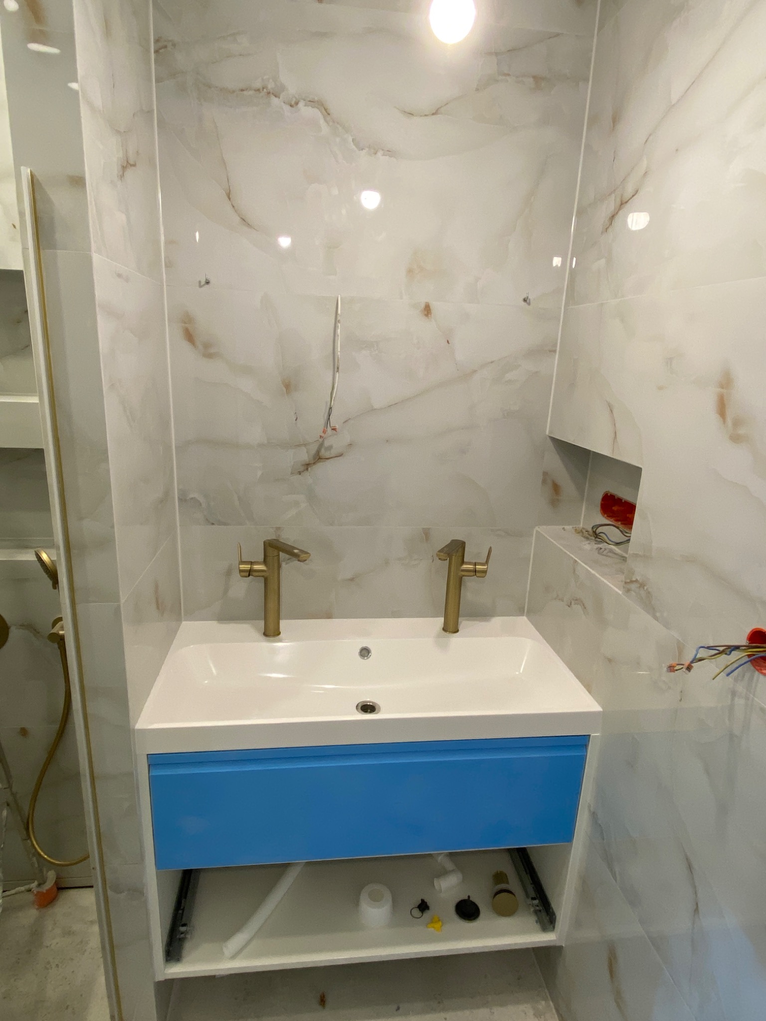 Łazienka w trakcie wykańczania: biała umywalka z niebieską szafką, złote baterie, marmurowe płytki na ścianach, widoczne przewody elektryczne i elementy instalacji hydraulicznej.