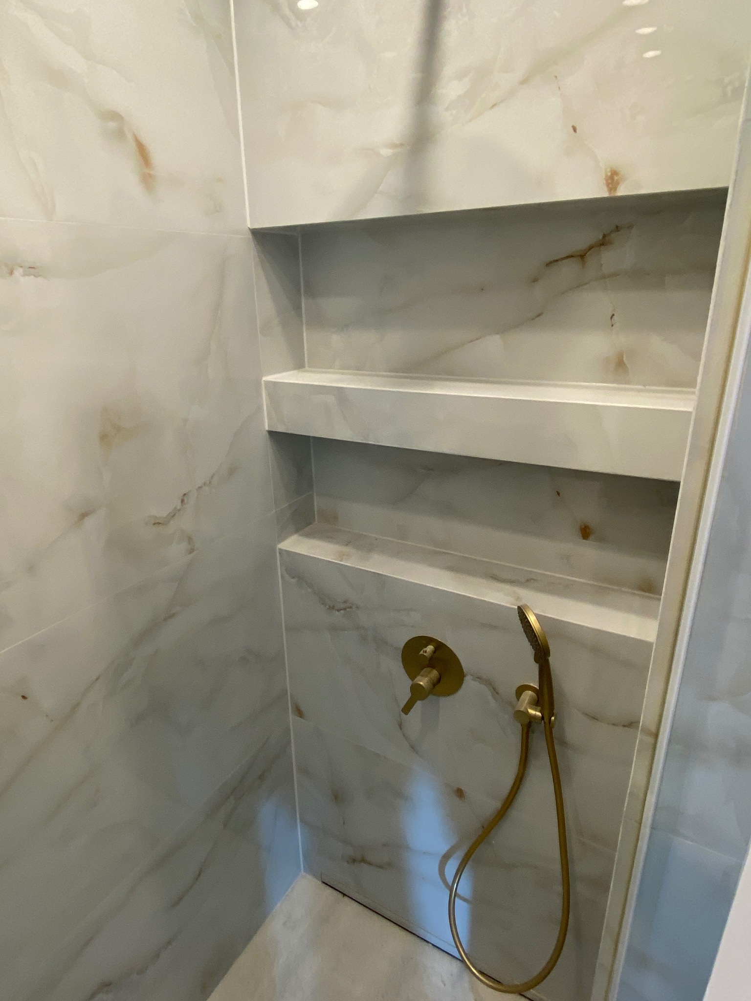 Wykończona kabina prysznicowa z półkami wnękowymi, wyłożona płytkami imitującymi marmur w odcieniach bieli i beżu, z zamontowaną złotą baterią i słuchawką prysznicową.