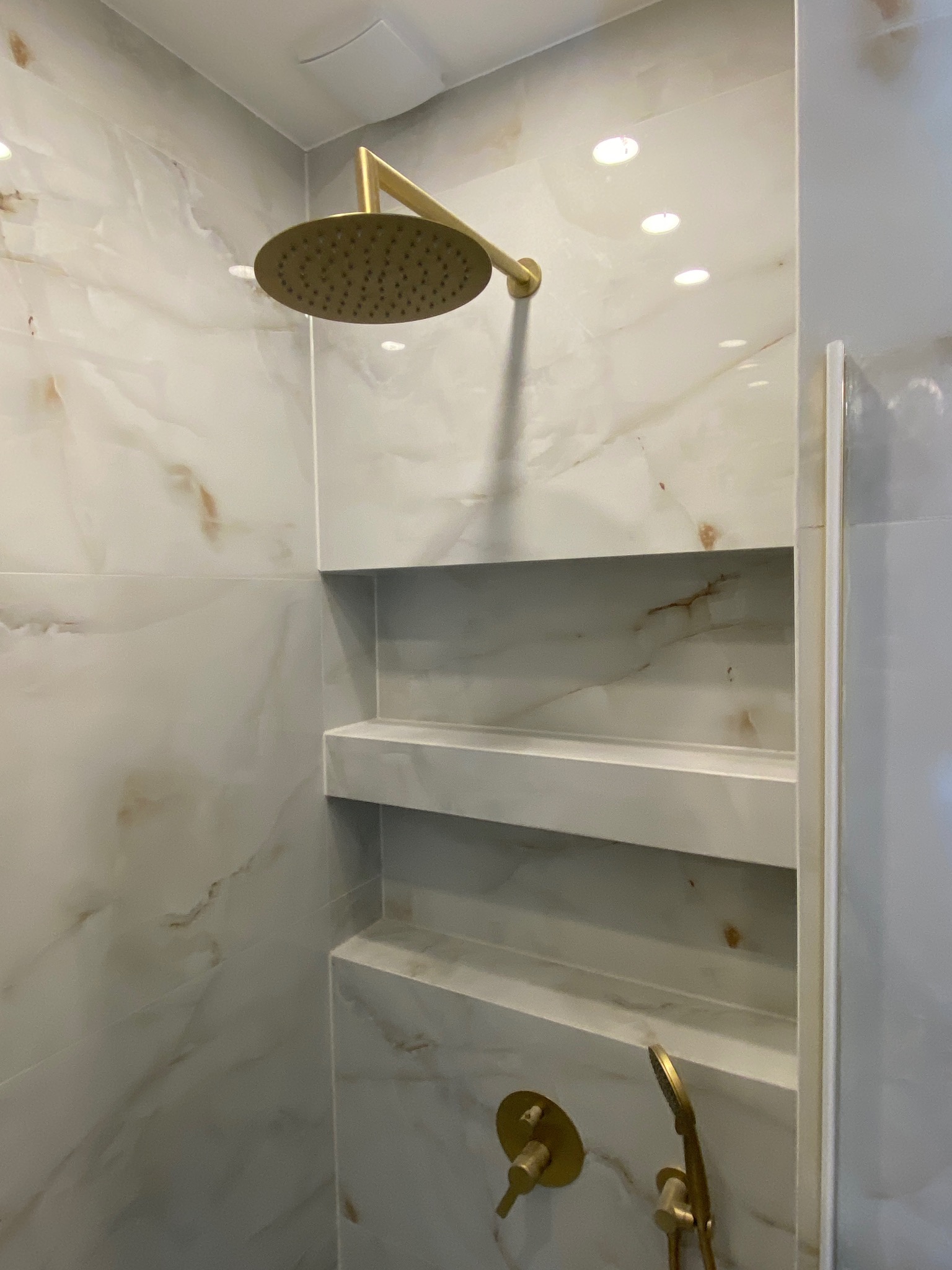 Wykończona kabina prysznicowa z marmurowymi płytkami, wbudowanymi półkami i złotą armaturą, oświetlona punktowymi lampami sufitowymi.