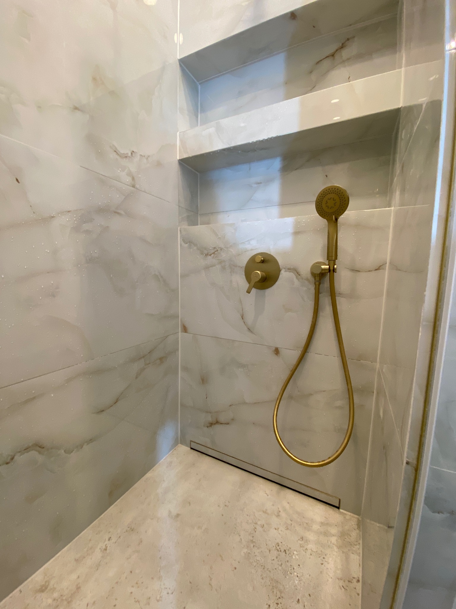 Kabina prysznicowa w Szczecinie wykończona marmurowymi płytkami z wnęką na kosmetyki i złotą armaturą, widoczne krople wody na ścianach.