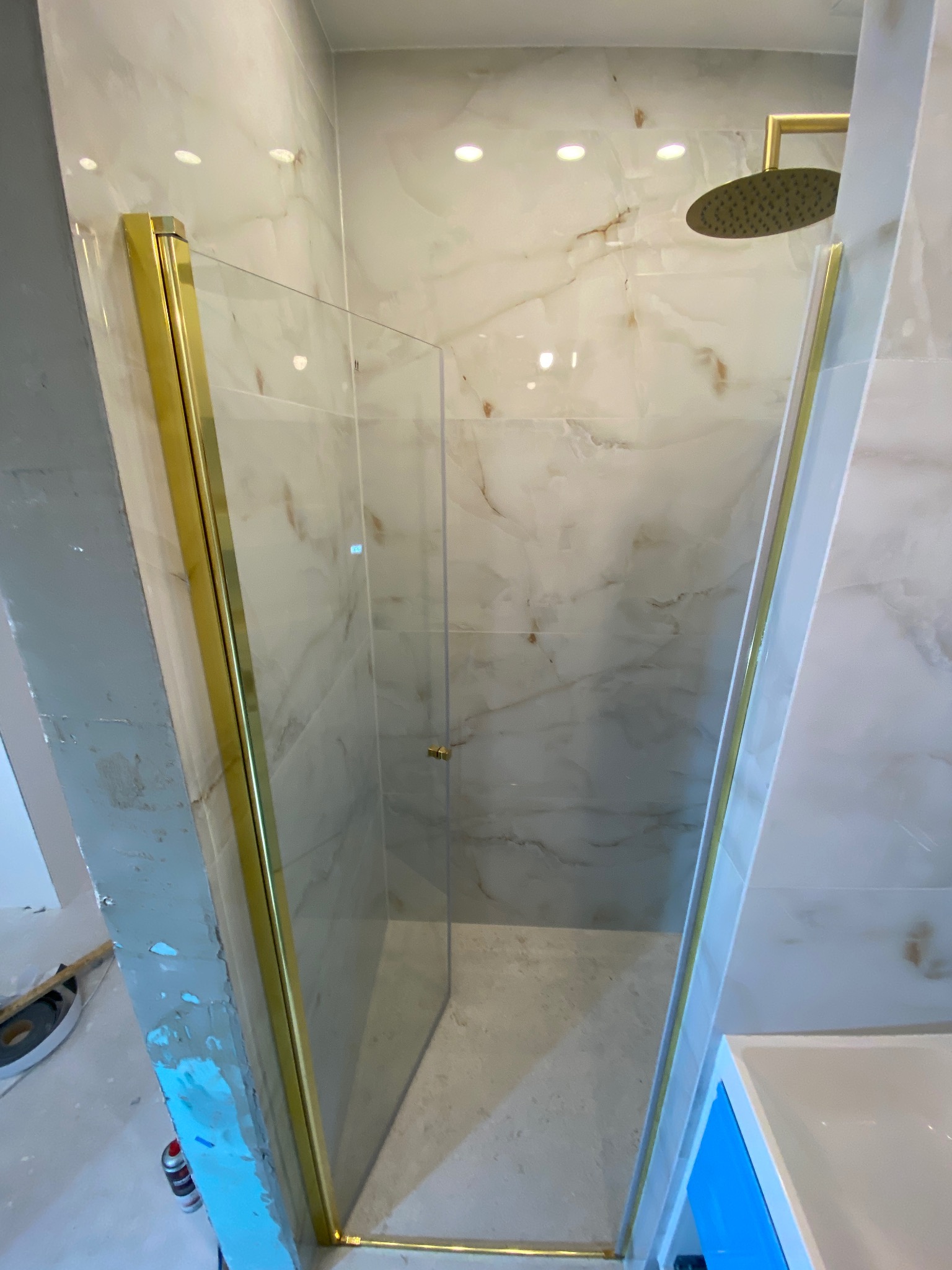 Szklana kabina prysznicowa z brokatowymi złotymi ramami, zamontowana w łazience z marmurowymi płytkami, widoczna słuchawka prysznicowa, częściowo wykończone ściany obok kabiny z widocznymi...