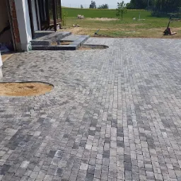 Ułożona szara kostka brukowa przed domem z wyznaczonym miejscem na rabatę, widoczny wózek paletowy w tle.