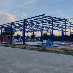 Wznoszenie szkieletu hali stalowej pomalowanego na niebiesko, widoczny fundament i częściowo wymurowana ściana z czerwonej cegły. W tle zaparkowane samochody ciężarowe.