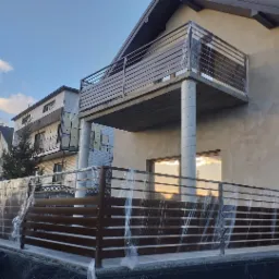 Nowoczesny dom z balkonem zabezpieczonym poziomą balustradą oraz ogrodzenie posesji z poziomych profili, oba zabezpieczone folią ochronną na czas prac wykończeniowych elewacji.