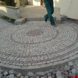 Okrągły wzór z jasnoszarej i brązowej kostki na podjeździe, z osobą w zielonych spodniach wchodzącą na niego.