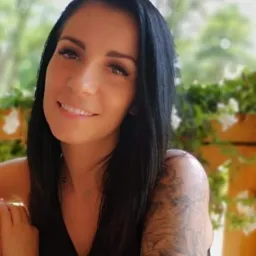 Uśmiechnięta kobieta z ciemnymi włosami i tatuażem na ramieniu, w tle zieleń