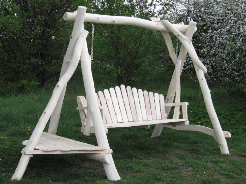 Drewniana huśtawka ogrodowa z siedziskiem wykonanym z pionowych listew, zawieszona na łańcuchach, z dodatkową półką z boku, wykonana z surowego drewna.