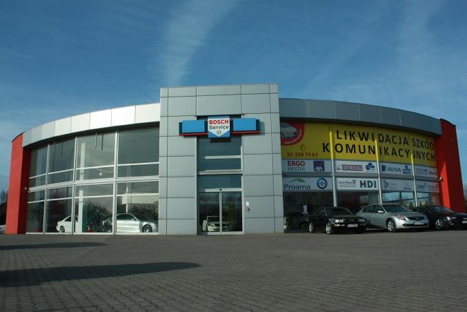 Budynek serwisu samochodowego Bosch Service z dużymi oknami wystawowymi i reklamami ubezpieczycieli, przed budynkiem zaparkowane samochody klientów.