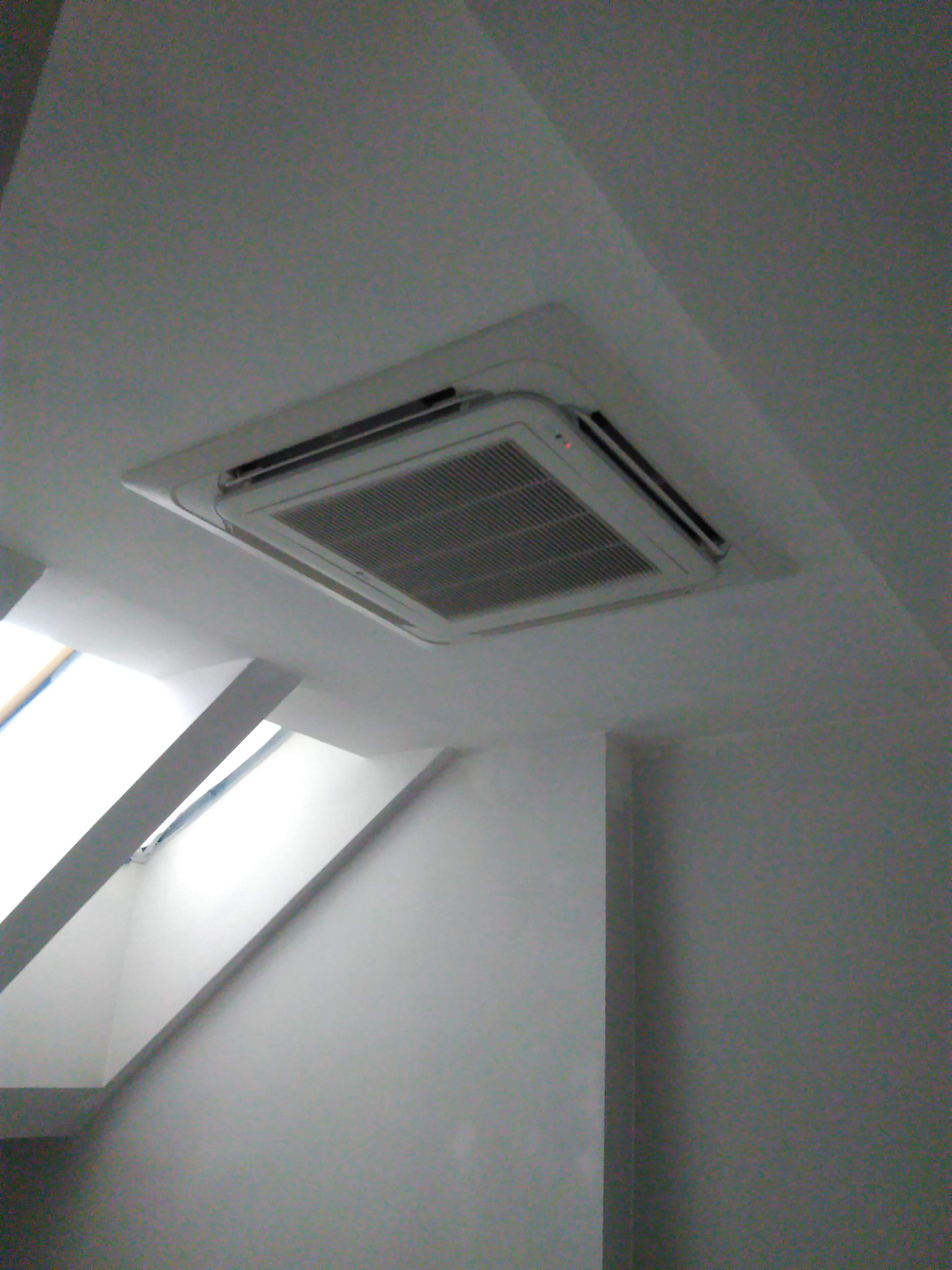 Kasetonowa jednostka wewnętrzna klimatyzacji zamontowana w suficie poddasza, widoczne okno dachowe w tle.