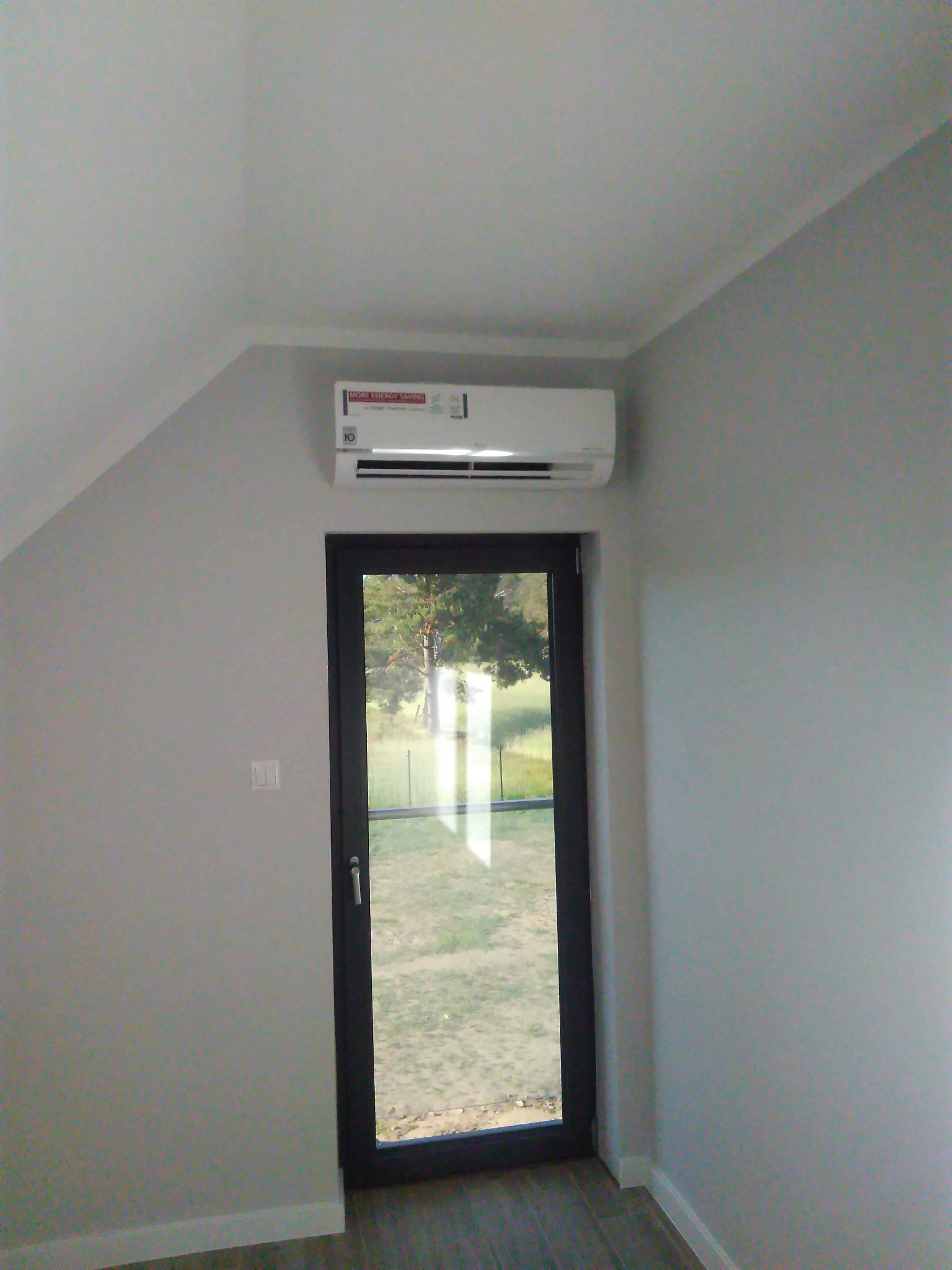 Biała jednostka klimatyzacji LG zamontowana nad czarnymi drzwiami balkonowymi w szarym pokoju na poddaszu.