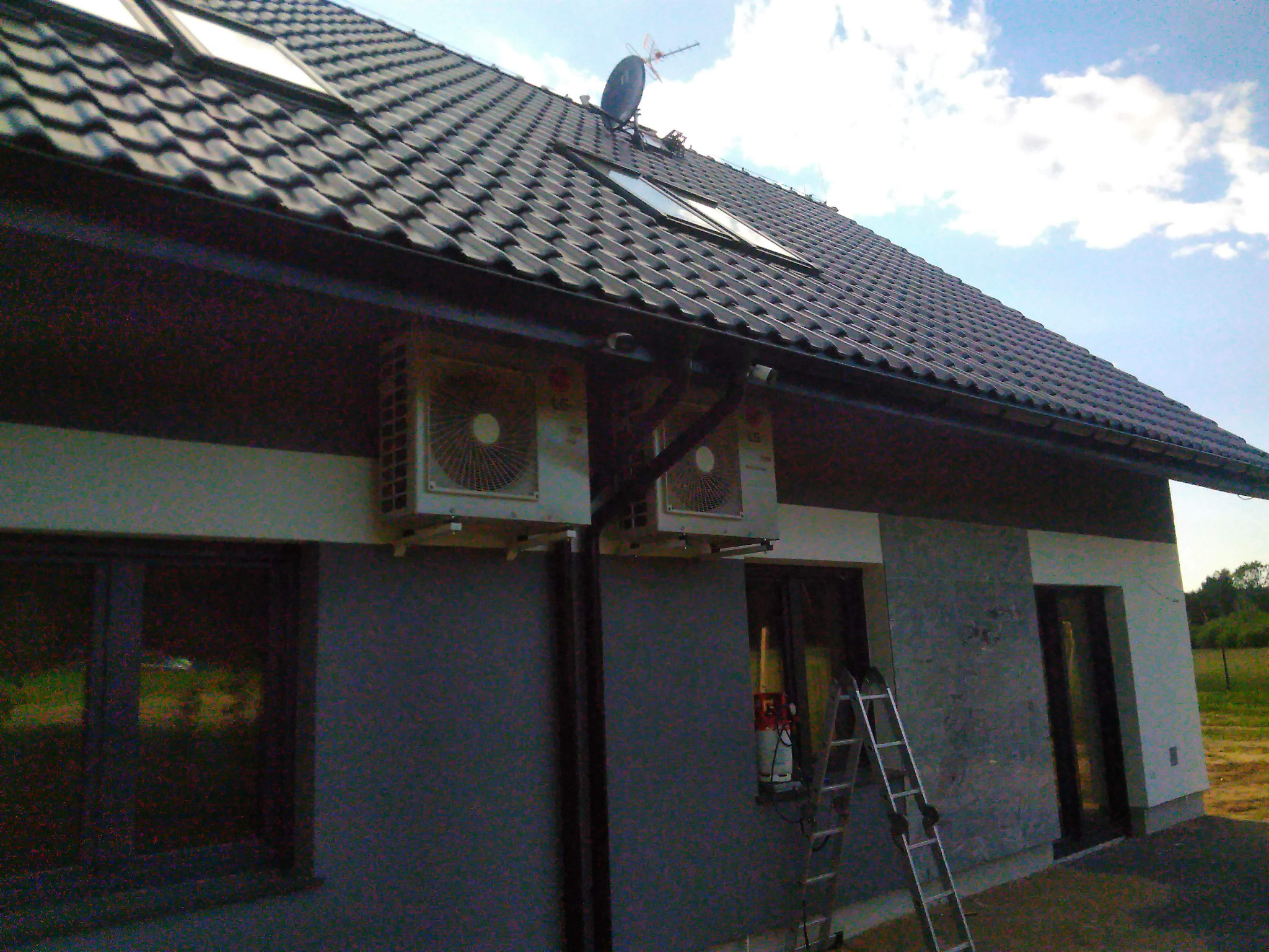 Dwa zewnętrzne moduły klimatyzacji zamontowane na elewacji domu z ciemną dachówką, widoczne okna dachowe, drabina oparta o ścianę, częściowo pochmurne niebo.