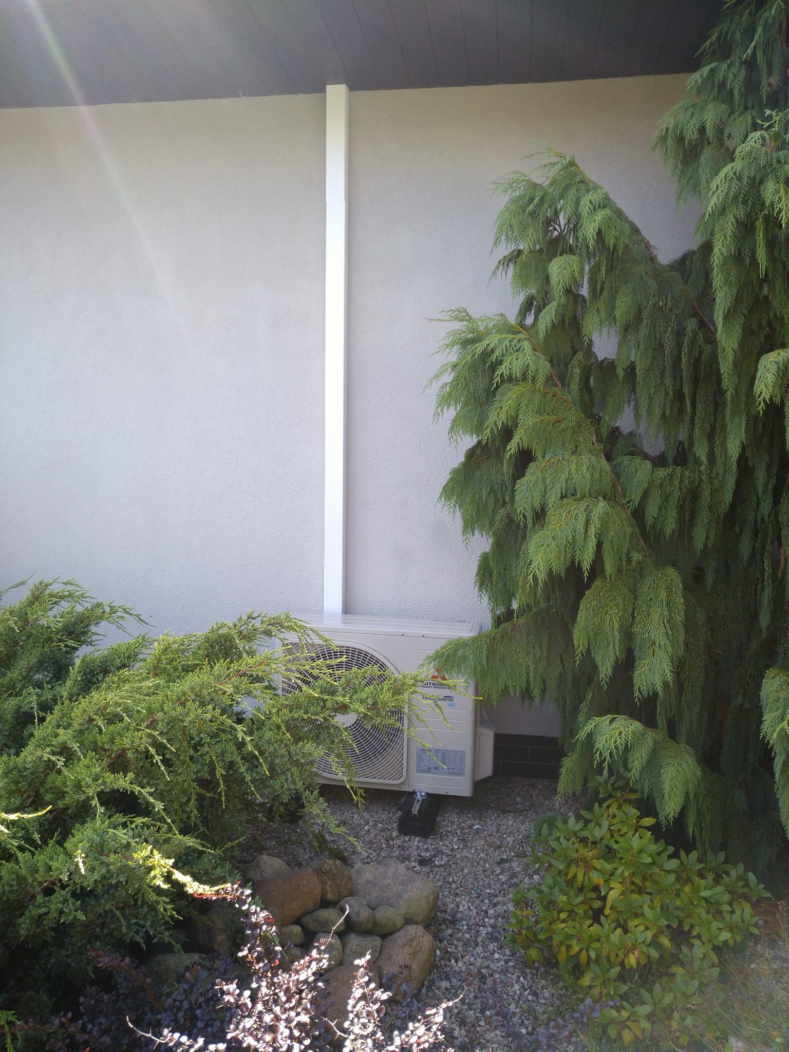 Zewnętrzna jednostka klimatyzacji Mitsubishi ukryta wśród zieleni krzewów iglastych i kamieni na tle jasnej ściany budynku, z widoczną białą rurą odprowadzającą.