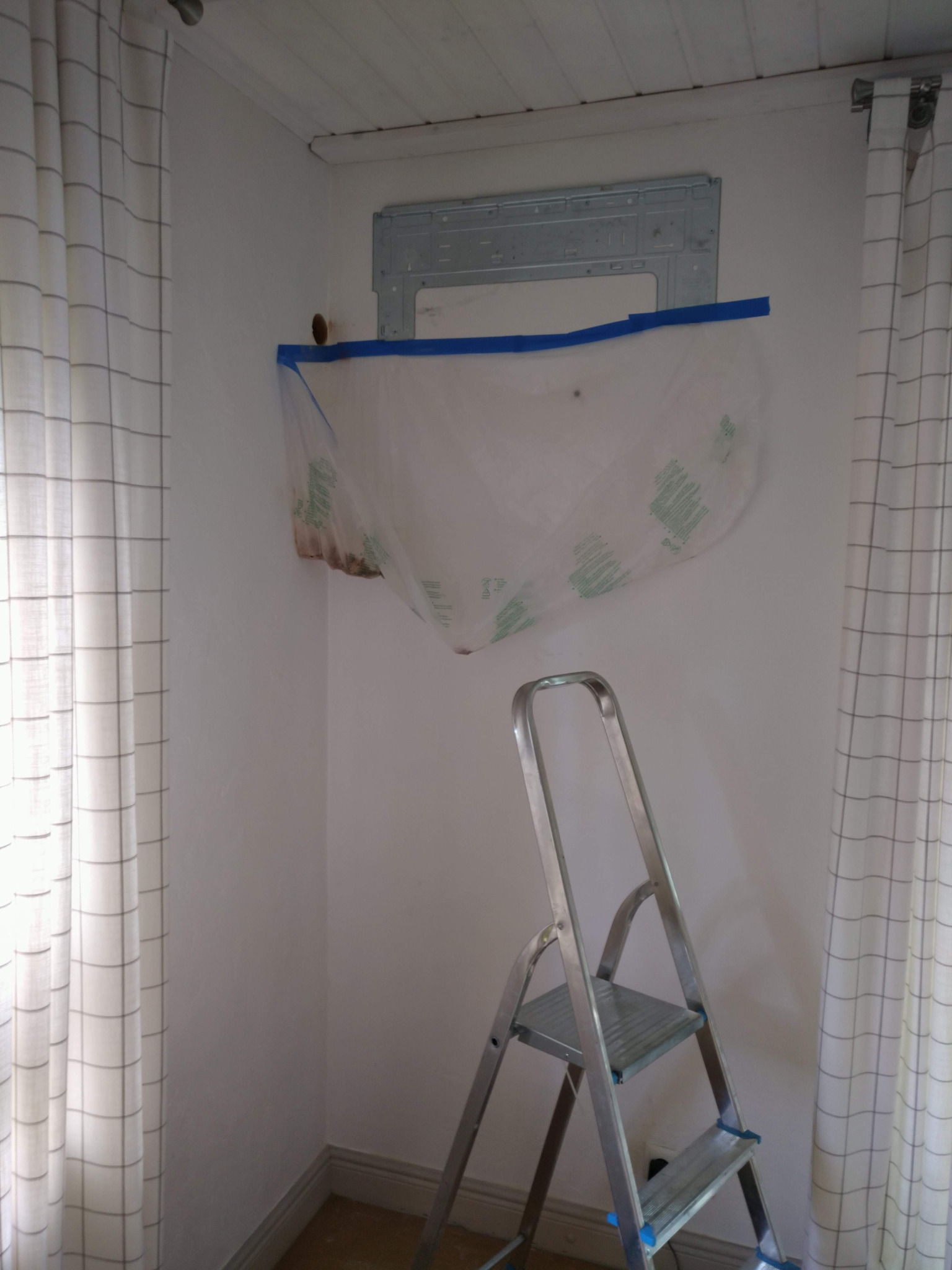 Przygotowanie do montażu klimatyzacji: zamaskowany otwór w ścianie, zabezpieczony folią i taśmą malarską, drabina aluminiowa w tle, zasłony w kratę po bokach.