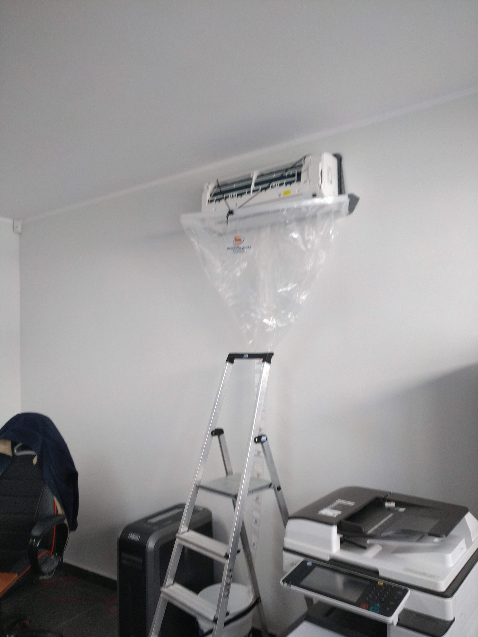 Wnętrze pokoju z klimatyzatorem na ścianie zabezpieczonym folią ochronną podczas czyszczenia, obok drabina aluminiowa, niszczarka i drukarka wielofunkcyjna.