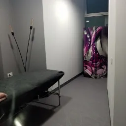 Wnętrze gabinetu z łóżkiem do zabiegów, lampami podłogowymi i dekoracyjnym parawanem. Szara kolorystyka pomieszczenia.