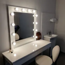 Biała toaletka z lustrem otoczonym żarówkami, obok umywalka z szafką i dekoracyjna rzeźba drzewa życia. Na blacie toaletki małe ozdoby.