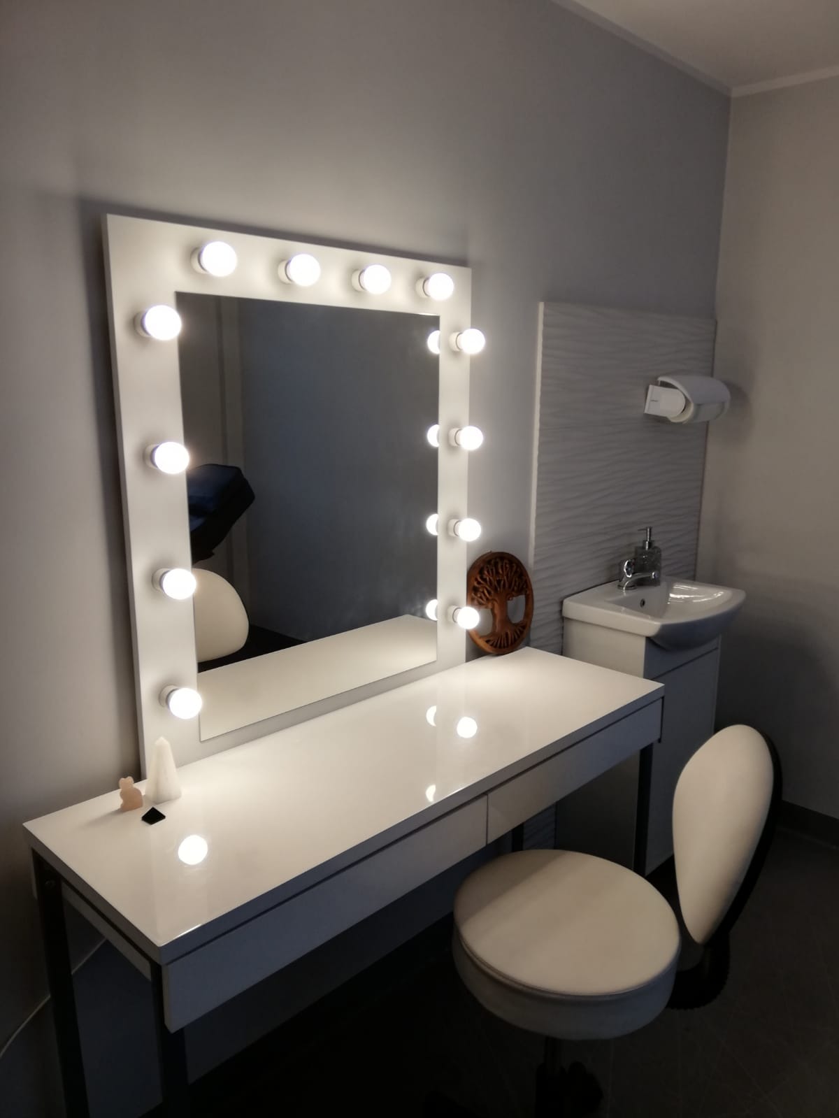 Biała toaletka z lustrem otoczonym żarówkami, obok umywalka z szafką i dekoracyjna rzeźba drzewa życia. Na blacie toaletki małe ozdoby.