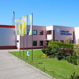 Budynek zakładu stolarskiego z widocznym logo firmy i flagami reklamowymi przed wejściem, trawnik z ozdobnymi krzewami, brama garażowa po lewej stronie.