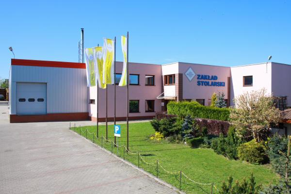 Budynek zakładu stolarskiego z widocznym logo firmy i flagami reklamowymi przed wejściem, trawnik z ozdobnymi krzewami, brama garażowa po lewej stronie.