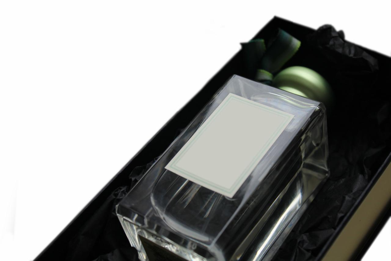 Elegancki flakon perfum z pustą etykietą w czarnym, teksturowanym pudełku prezentowym, widoczne zielone akcenty dekoracyjne.