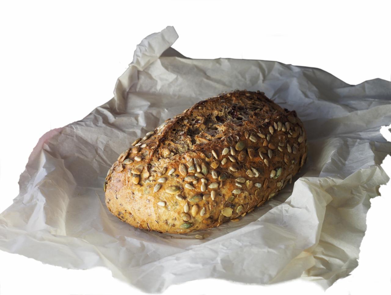 Bochenek rustykalnego chleba posypanego ziarnami dyni, ułożony na pogniecionym, cienkim papierze.