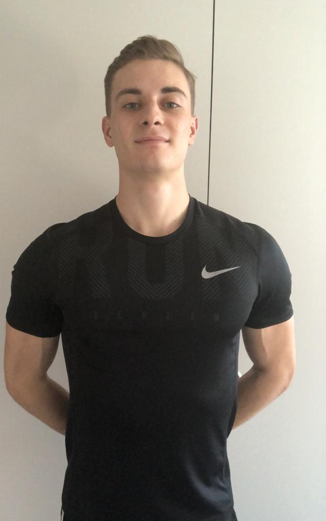 Młody mężczyzna w czarnej koszulce sportowej z logo Nike, pozujący na tle jasnej ściany.