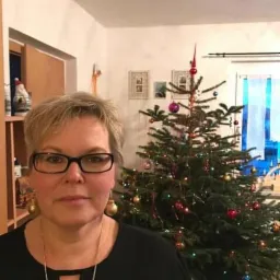 Portret kobiety w okularach na tle choinki z bombkami i światełkami. Kobieta ma krótkie blond włosy i czarną bluzkę. W tle widoczne dekoracje świąteczne.