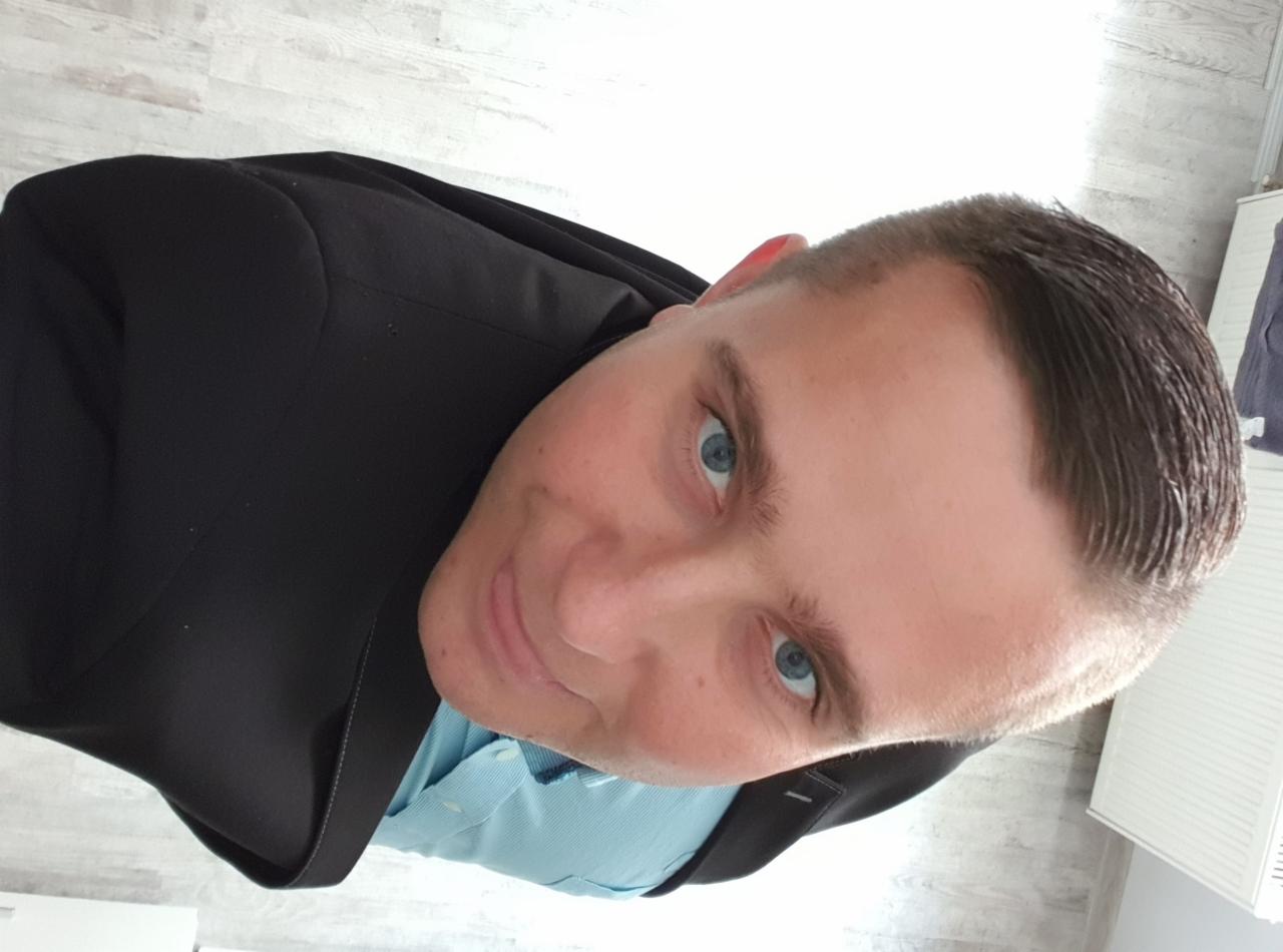 Portret mężczyzny w garniturze, z niebieskimi oczami, zrobiony z perspektywy żabiej w Bolesławicach. Uśmiechnięta twarz i krótko ostrzyżone włosy.