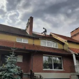 Pracownik w uprzęży na dwuspadowym dachu z czerwonej dachówki, czyści go pod ciśnieniem, widoczny ceglany komin i fragment elewacji domu z czerwonej cegły na tle pochmurnego nieba.