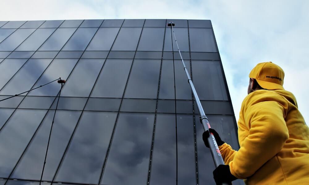 Pracownik w żółtej bluzie i czarnych rękawicach czyści szklaną fasadę biurowca za pomocą teleskopowej lancy z dyszą, widoczne odbicie chmur w szybach.