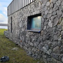 Fragment ściany budynku wykończonej kamieniem naturalnym, z widocznym oknem i fragmentem metalowej elewacji na górze, z młotkiem murarskim leżącym u podstawy ściany.