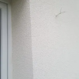 Fragment białej elewacji z fakturą baranka, narożnik okienny i przyklejony patyczek w kształcie litery Y.