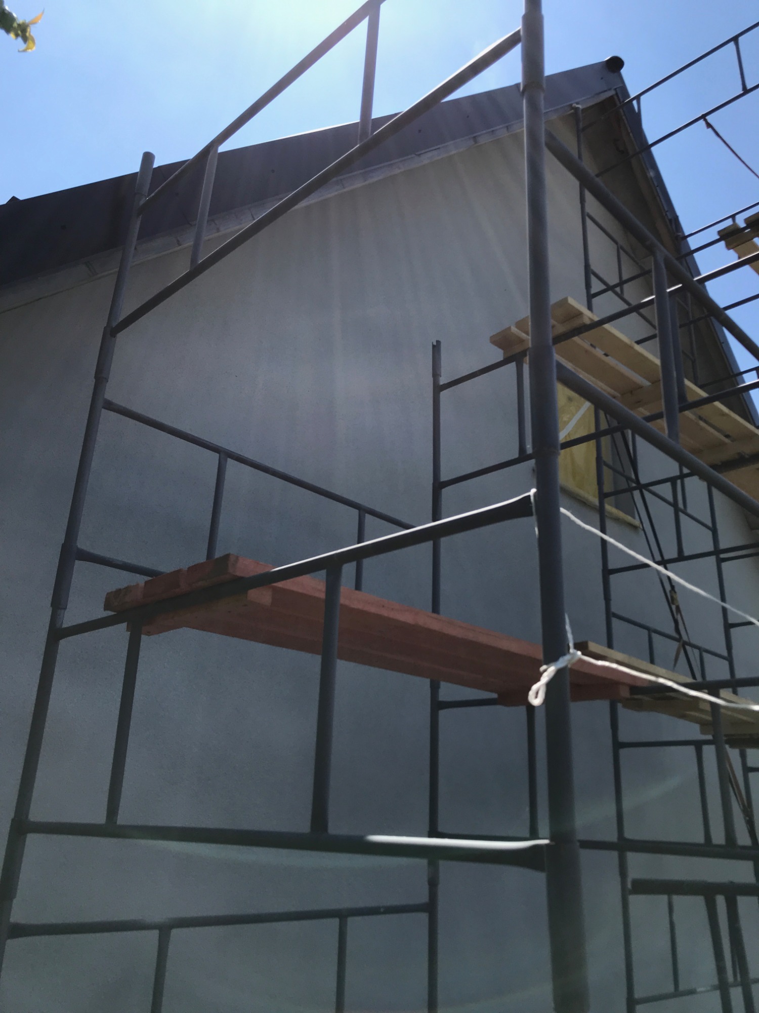 Szarego koloru elewacja budynku w trakcie prac wykończeniowych z widocznym rusztowaniem i deskami.