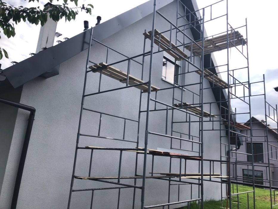 Dom jednorodzinny z szarą elewacją w trakcie tynkowania, widoczne rusztowania z drewnianymi pomostami i fragment dachu z kominem.
