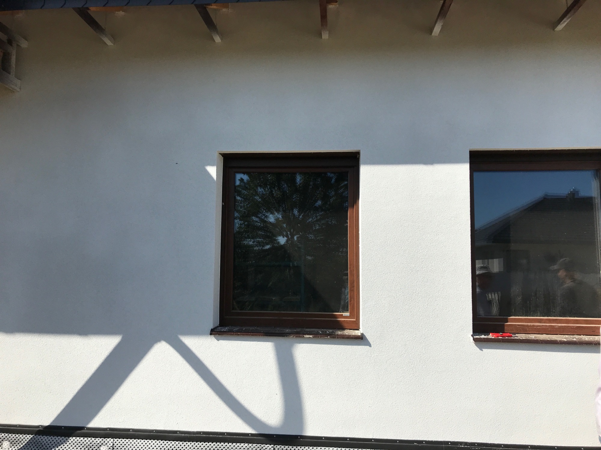 Biała elewacja budynku z dwoma oknami o brązowych ramach, na parapecie jednego z okien leżą czerwone narzędzia, widoczne cienie rzucane przez elementy konstrukcyjne dachu.