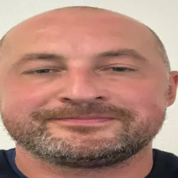 Portret mężczyzny z krótkimi włosami i brodą, na jednolitym tle.