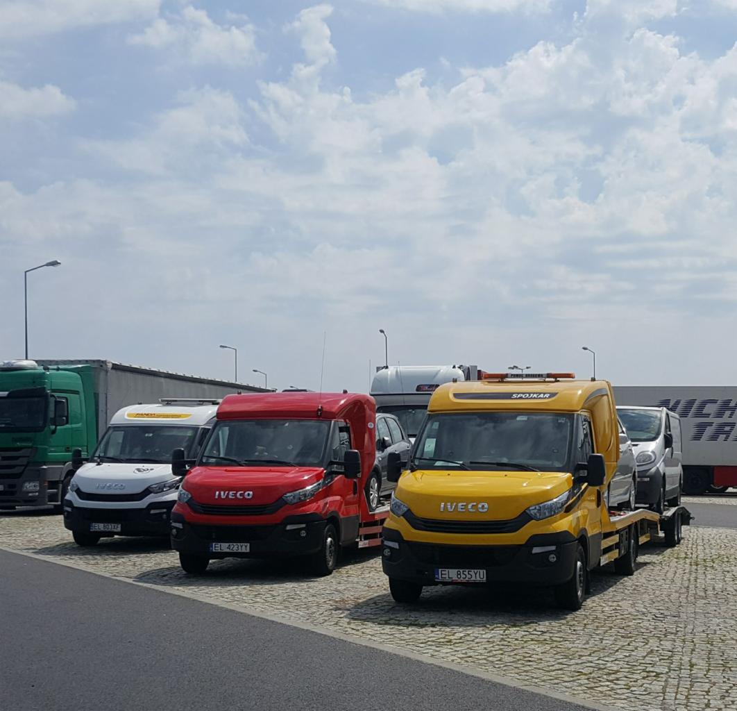 Żółta laweta Iveco z załadowanym samochodem osobowym, zaparkowana na placu obok czerwonej i białej lawety, w tle inne pojazdy i błękitne niebo z chmurami.