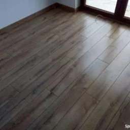 Ułożone panele podłogowe w odcieniach brązu, widoczne przy ścianie i oknie, z delikatnym połyskiem i strukturą drewna.