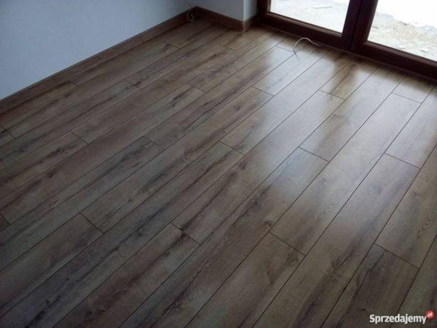 Ułożone panele podłogowe w odcieniach brązu, widoczne przy ścianie i oknie, z delikatnym połyskiem i strukturą drewna.