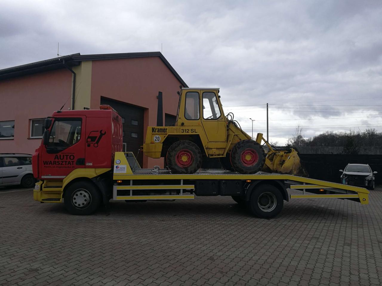 Żółta ładowarka Kramer 312 SL na czerwono-żółtej lawecie Auto Warsztat na tle budynku z szarymi drzwiami garażowymi i pochmurnego nieba.