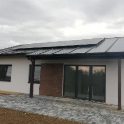 Nowoczesny dom jednorodzinny z ciemną blachą na dachu i panelami słonecznymi, wykończony szarą kostką brukową na podwórku, widok z boku w pochmurny dzień.