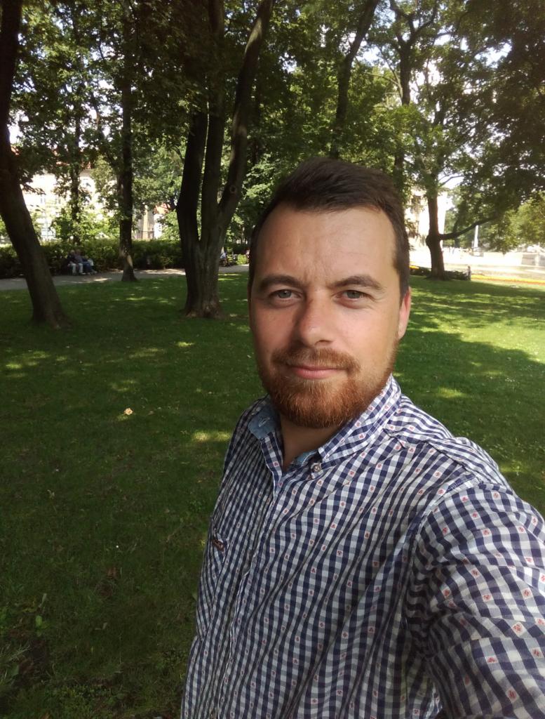 Mężczyzna w koszuli w kratę, selfie w parku, zielona trawa, drzewa w tle. Uśmiechnięty, z brodą. Letnie światło, naturalne otoczenie.