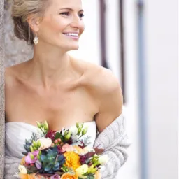 Uśmiechnięta panna młoda w sukni bez ramiączek, z upiętymi blond włosami i kolczykami, trzyma kolorowy bukiet kwiatów, ubrana w sweter.