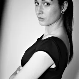 Portret kobiety w czarnej sukience, z włosami związanymi w kucyk, spoglądającej przez ramię na czarno-białym zdjęciu studyjnym.