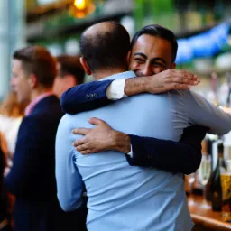 Uśmiechnięty mężczyzna w błękitnej koszuli obejmuje innego mężczyznę w ciemnej marynarce na tle baru z butelkami alkoholu i rozmytych postaci gości.