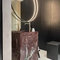 Nowoczesna łazienka z umywalką z czerwonego marmuru, okrągłym lustrem z podświetleniem LED i ciemnymi drewnianymi panelami na ścianach.