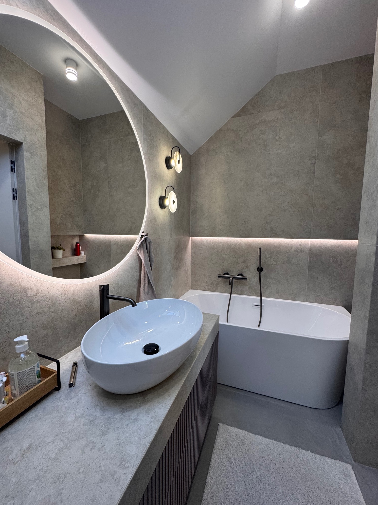 Nowoczesna łazienka z owalną umywalką, podświetlanym lustrem i wanną. Szare płytki, minimalistyczne oświetlenie i akcenty w kolorze czarnym tworzą elegancki wystrój.