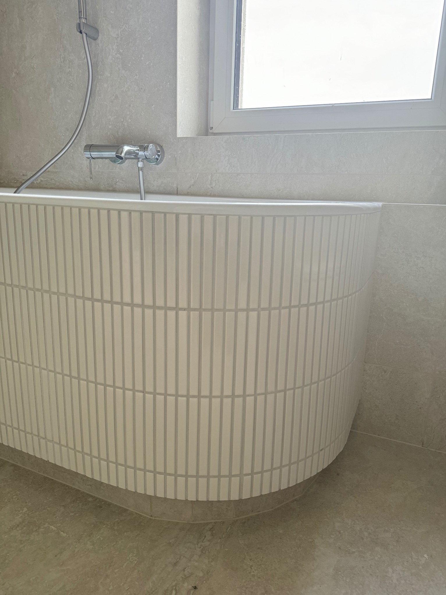 Biała wanna obłożona pionowymi, prostokątnymi płytkami w łazience z szarą armaturą i oknem. Minimalistyczny design, jasne wnętrze.